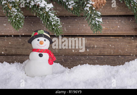 Pupazzo di neve vacanze figurina sulla neve con evergreen banches su uno sfondo di legno con la caduta della neve Foto Stock
