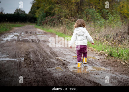 Il Toddler girl, vista posteriore, spruzzi in pozze fangose in strada di campagna con giallo stivali da pioggia. Copia di sinistra dello spazio. Foto Stock