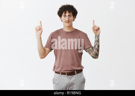 Grave-alla moda ed elegante guy con eleganti baffi capelli ricci e tatuaggi sui bracci di sollevamento mani rivolte verso l'alto e guardando con calma spensierata exp Foto Stock