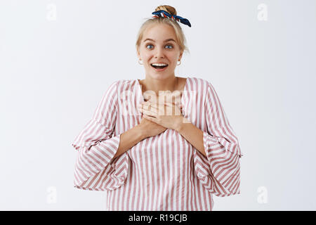 È possibile effettuare il cuore batte più veloce. Ritratto di incantato e sorpreso carino donna moderna in camicia a righe e fascia, sorridente largamente, tenendo le palme sul petto una Foto Stock