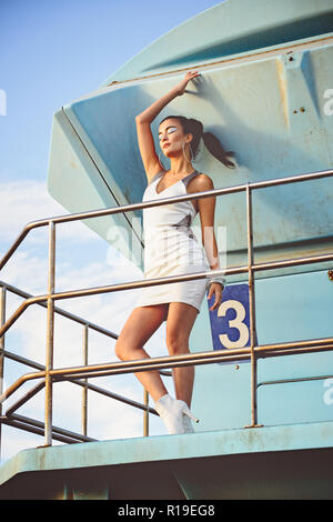Una bella asiatica modello femminile, razza mista, in posa su un bagnino post, un esterno di moda concetto editoriale. Foto Stock