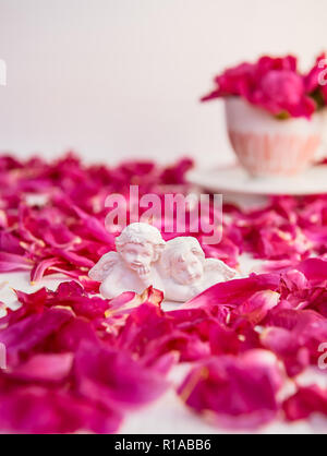 Statuetta di due antichi piccoli angeli incantevole del gesso su sfondo bianco con rosa peonia viola petali di fiori. Amore, romantico, Valentines d Foto Stock