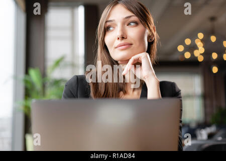 Giovane donna dietro lo schermo del laptop. premurosamente guardare lontano. Ragazza blogging, navigare in Internet e chattare. Si siede in un coffee shop con ampia finestra. Foto Stock