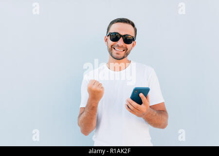 Giovane uomo barbuto con vittoria urlare guardando il messaggio mobile a sfondo blu. Foto Stock