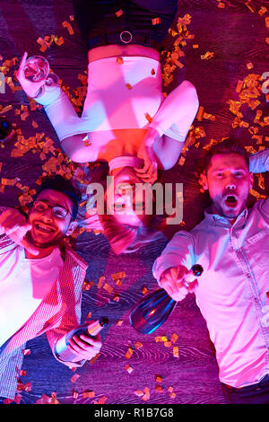 Da sopra il colpo di ridere gli uomini e una donna con champagne giacente sulla farina in neon luci aventi la festa di Capodanno Foto Stock