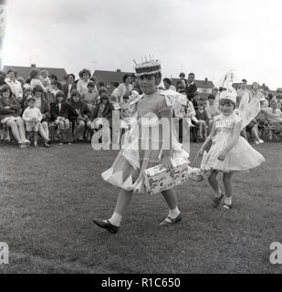 1967, historcial, al di fuori di un fieldd, in un villaggio inglese fete, due giovani ragazze vestito in costume a piedi di fronte gli spettatori in un abito di fantasia concorrenza, Inghilterra, Regno Unito. Una delle ragazze ha un partito vestito su e indossando una torta sul suo capo, mentre l altro è vestito come un angelo e ha parole Gypsy Moth IV su di lei il nome della barca di Sir Francis Chichester che il mese seguente, correttamente navigato, con una sola mano, in tutto il mondo. I suoi pensieri erano con lui. Foto Stock