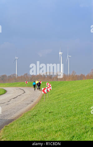 Tre piloti in bicicletta sulla strada di un paese e delle turbine a vento sul cielo blu in background Foto Stock