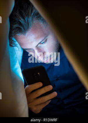 Giovane uomo nascondendosi sotto una coperta mentre si utilizza un telefono cellulare per la navigazione in internet o per chattare privatamente online Foto Stock