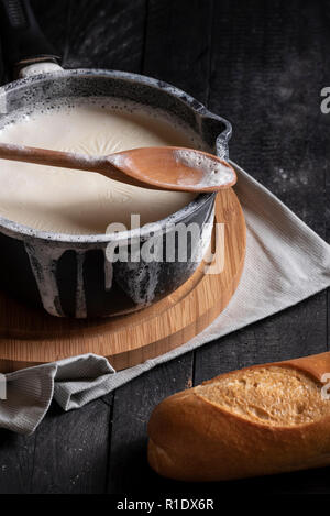 Latte bollito in una pentola nera e rovesciato sopra i bordi, su di un tagliere di legno e un asciugamano da cucina, ed un pane francese, su un tavolo rustico. Foto Stock