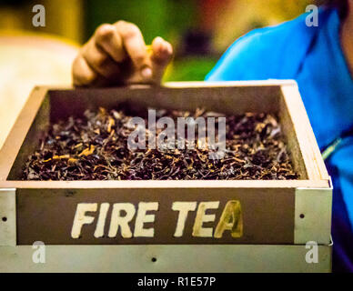 Una mano prende un campione di tè nero appena tostato da una scatola di legno etichettata "Fire Tea" alla Blue Field Tea Estate a Mount Harrow vicino a Ramboda, vicino a Nuwara Eliya, Sri Lanka. La piantagione mostra ai visitatori l'intero processo di produzione del tè Ceylon qui, dall'appassimento al rullatura e alla fermentazione fino alla tostatura e al vagliamento. La tostatura utilizza il calore per fermare la fermentazione e asciugare la foglia fino al suo stato finale, conservabile. Foto Stock