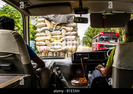 Un camion carico di sacchi avanza davanti all’autobus turistico nel traffico a sinistra, mentre un autobus interurbano rosso si avvicina dall’altro lato della strada vicino a Hambantota, Sri Lanka. La vista attraverso il parabrezza cattura il traffico intenso sulla strada da Colombo attraverso Galle a Hambantota. Foto Stock