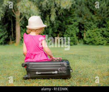 Vista dal retro, anonimi ragazza giovane bambino in abito rosa si siede su un nero per i bagagli a mano e attende, parco all'aperto in estate. Foto Stock