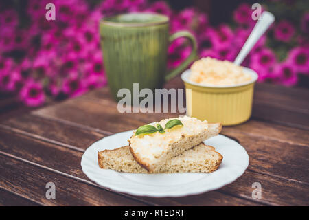 Giallo delizioso uovo e maionese sulla pasta di pane integrale, prima colazione snack ricetta, fiori di colore rosa sullo sfondo, base in legno. Foto Stock