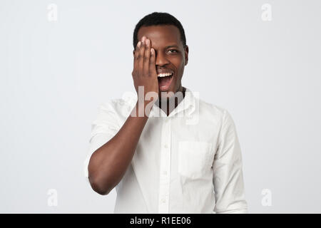 I giovani africani Uomo in camicia bianca che copre metà faccia con la mano mentre in piedi contro uno sfondo grigio. Positivo emozione umana Foto Stock