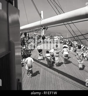 Degli anni Cinquanta, storico in mare a bordo di un' unione-castello steamship, femmina i passeggeri che prendono parte a una sacca gara fuori sul ponte della nave applauditi da guardare gli spettatori, molti seduti in sedie a sdraio. Foto Stock