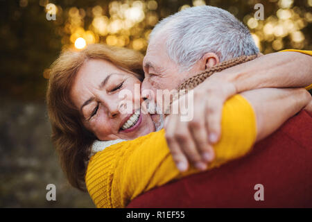 Una coppia senior in piedi in un autunno natura al tramonto, avvolgente e baciare. Foto Stock