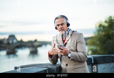 Imprenditore maturo con le cuffie e lo smartphone in piedi dal fiume, cantando. Foto Stock