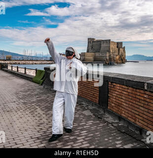 Pulcinella tradizionale maschera napoletana gesti con il Castel dell'Ovo in background nel Golfo di Napoli. Foto Stock