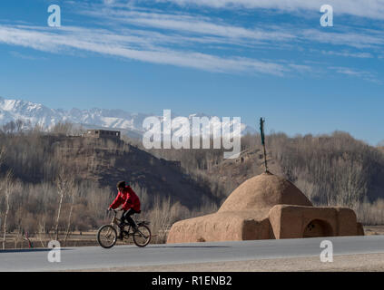 Ragazzo ciclismo su strada asfaltata entrando nella valle di Bamyan con un santuario e Snow-Capped montagne sullo sfondo, nella provincia di Mazar-i-Sharif, Afghanistan Foto Stock