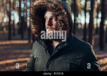 Ritratto, close-up di giovani gravi elegantemente con barba vestito in rgreen giacca invernale con un cappuccio e pelliccia sulla sua testa si erge contro il retro Foto Stock