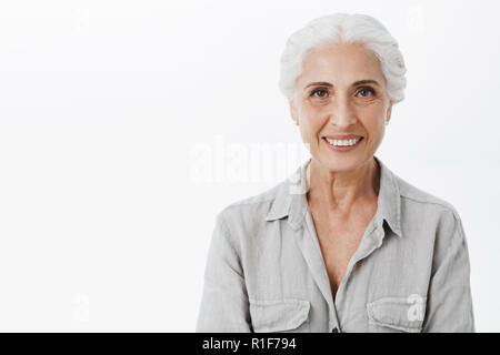 Vita-up shot di carino e gentile saggia donna anziana con i capelli bianchi in camicia casual sorridente largamente con assicurato e piacere guardare essendo divertito e carismatico in posa contro uno sfondo grigio Foto Stock