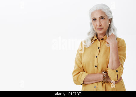 Studio shot di elegante e femminile donna anziana con i capelli bianchi orecchini perla e giallo alla moda di trench coat in posa contro uno sfondo grigio calmo e sicuro Foto Stock