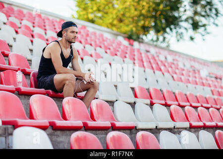 Grave uomo sportivo in ascolto in cuffia la musica seduto sulla tribuna dello stadio sedi Foto Stock