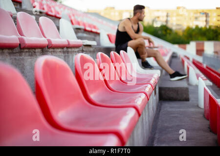 Righe di posti vuoti in un sport stadium, giovane sportivo sullo sfondo. Rosso e sedie in plastica bianca in attesa per i tifosi Foto Stock