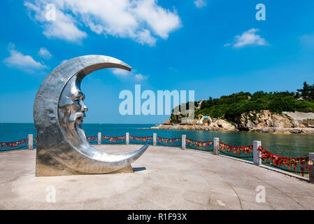 YANTAI, Shandong, Cina - 16LUG2018: Luna scultura a Moon Bay, con l'Est Fort Dongpaotai area dietro Foto Stock