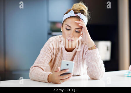 Stanco donna millenario appoggiato la testa in cucina e utilizza lo smartphone Foto Stock