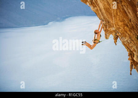 Femmina di rocciatore saltando sulle maniglie sul tracciato impegnativo sulla scogliera con vista sul mare e sulla costa di seguito Foto Stock