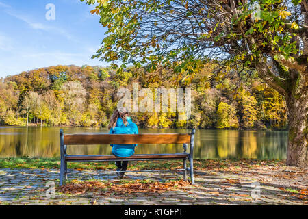 Vista posteriore di una ragazza su una panchina in riva al lago in autunno, Ulmen Maar, West Eifel campo vulcanico, regione della Renania, Germania, Europa Foto Stock