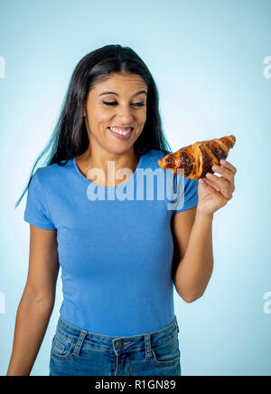 Giovane e bella carino e felice latino donna in abiti casual azienda big deliziosi croissant al cioccolato cercando con la tentazione di pensare se ignorando di Foto Stock