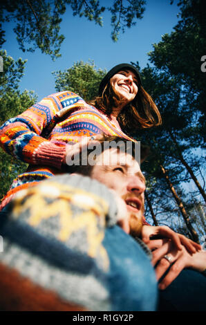 Gioiosa giovane ragazza si siede sul ragazzo spalle durante la passeggiata nella foresta, felice coppia indossando maglioni avendo divertimento all'aperto Foto Stock