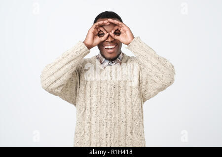 Bel giovane uomo africano, sorridente e rendendo il binocolo con le sue mani che indossa un maglione. Foto Stock