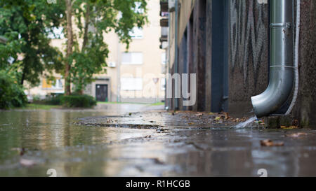 Tubo iniziale dello scarico acqua su un pattino della parte iniziale dello scarico direttamente in un invaso street. Poche gocce di pioggia sono che colpisce una superficie di una pozzanghera. Messa a fuoco selettiva. Foto Stock