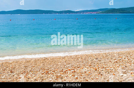 Ghiaiosa spiaggia di sabbia sullo sfondo di una collina gamma con molte piccole case, una linea di boe e azzurro trasparente mare blu. Blu cielo privo di nuvole. Zadar Foto Stock