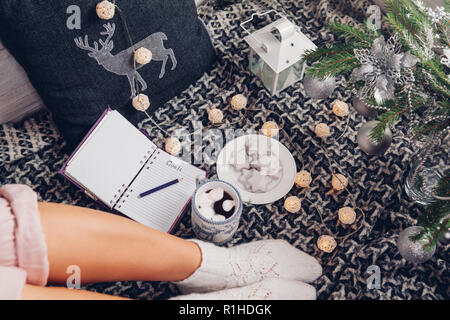 La scrittura di obiettivi per il prossimo anno con una tazza di cioccolato e biscotti sotto l albero di Natale. Sentimento donna accogliente a casa Foto Stock