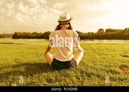 Giovane ragazza adolescente siede nel parco vicino al fiume, vista posteriore, ragazza guarda l'acqua, le pratiche yoga, medita. Foto Stock