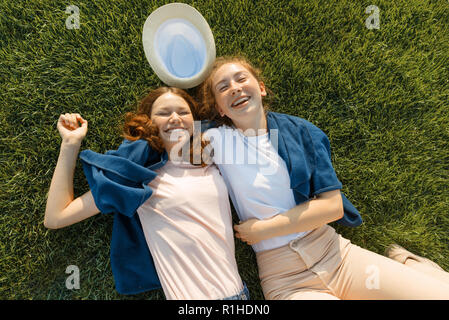 Due giovani ragazze sorridenti amici giacciono abbracciando sull'erba verde in estate soleggiato parco, vista dall'alto, le ragazze sono il divertimento in estate giornata di sole. Foto Stock