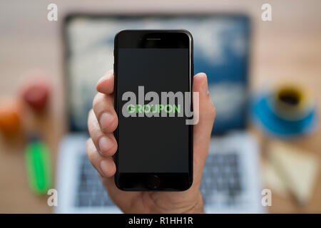 Un uomo guarda al suo iPhone che visualizza il logo Groupon, mentre se ne sta seduto alla sua scrivania per computer (solo uso editoriale). Foto Stock