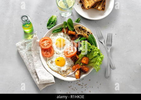 Vista da sopra il tavolo della colazione. La prima colazione la piastra con uova fritte, verdure, funghi e toast sul cemento grigio Sfondo. Sana colazione tabella concep Foto Stock