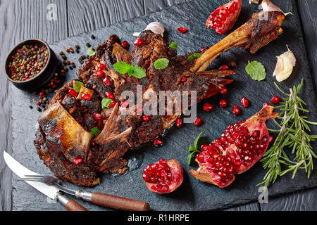 Deliziosa Spalla arrosto di agnello con i semi di melograno, foglie di menta, spezie e rosmarino su un nero ardesia piastra su una tavola di legno,vista da sopra, cl Foto Stock