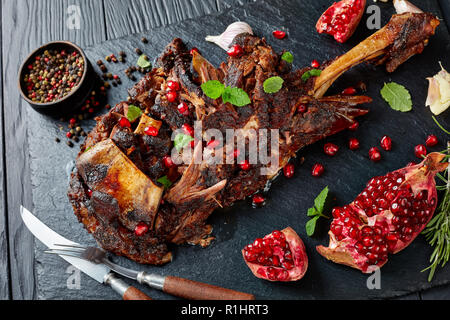 Vista aerea di deliziosi arrosti di gara la gamba di agnello serviti con i semi di melograno, foglie di menta, spezie e rosmarino su un nero ardesia piastra su un di legno Foto Stock