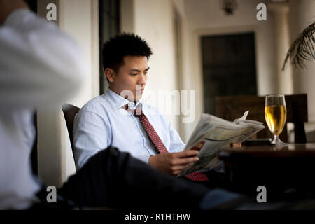 Giovane imprenditore leggendo un giornale seduti ad un tavolo con il suo collega e un bicchiere di birra. Foto Stock