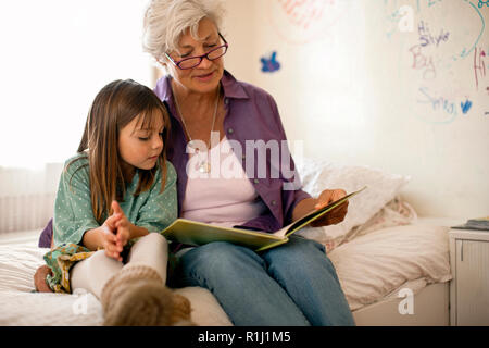 La nonna legge un libro con la sua giovane nipote come essi siedono insieme su un letto. Foto Stock