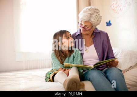 La nonna legge un libro con la sua giovane nipote come essi siedono insieme su un letto. Foto Stock