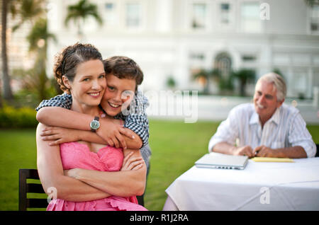 Ragazzo sorridente in piedi con le braccia intorno a sua madre, suo padre che guarda. Foto Stock
