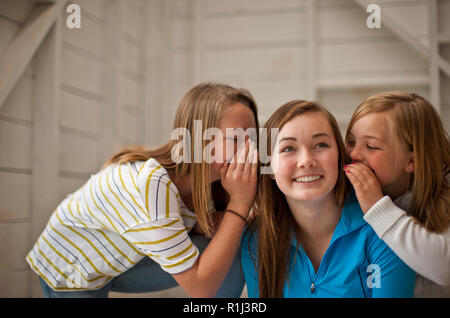 Grinning ragazza adolescente ascoltando le sue due giovani sorelle sussurro in entrambe le sue orecchie. Foto Stock