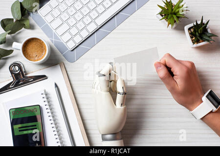 Immagine ritagliata di imprenditore con mano cyborg e smartwatch holding blank carta da visita a tavola con lo smartphone con prenotazione sullo schermo in office Foto Stock